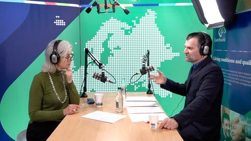 Two people wearing headphones sit at a table with microphones, discussing in a studio with a map backdrop.