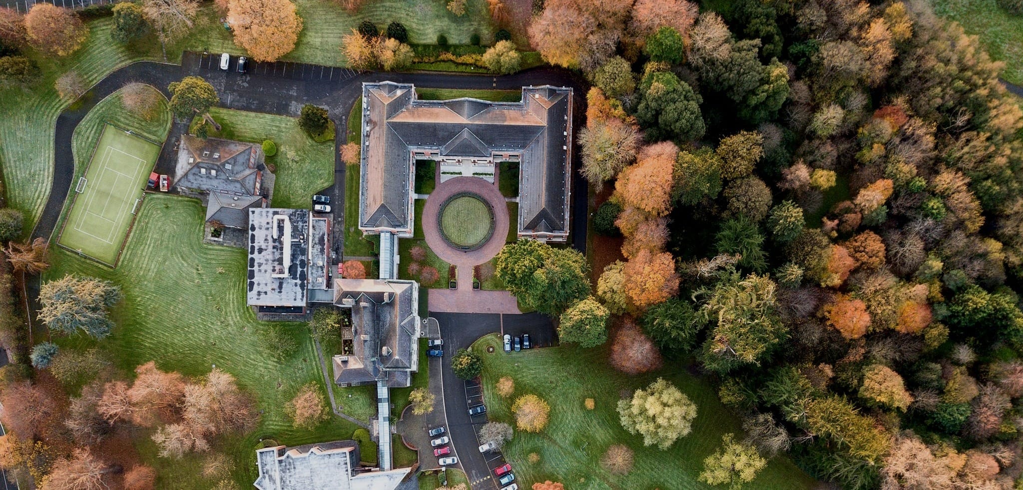 Aerial view of Eurofound premises with buildings, a circular courtyard, parking area, tennis court, and surrounding autumn trees.