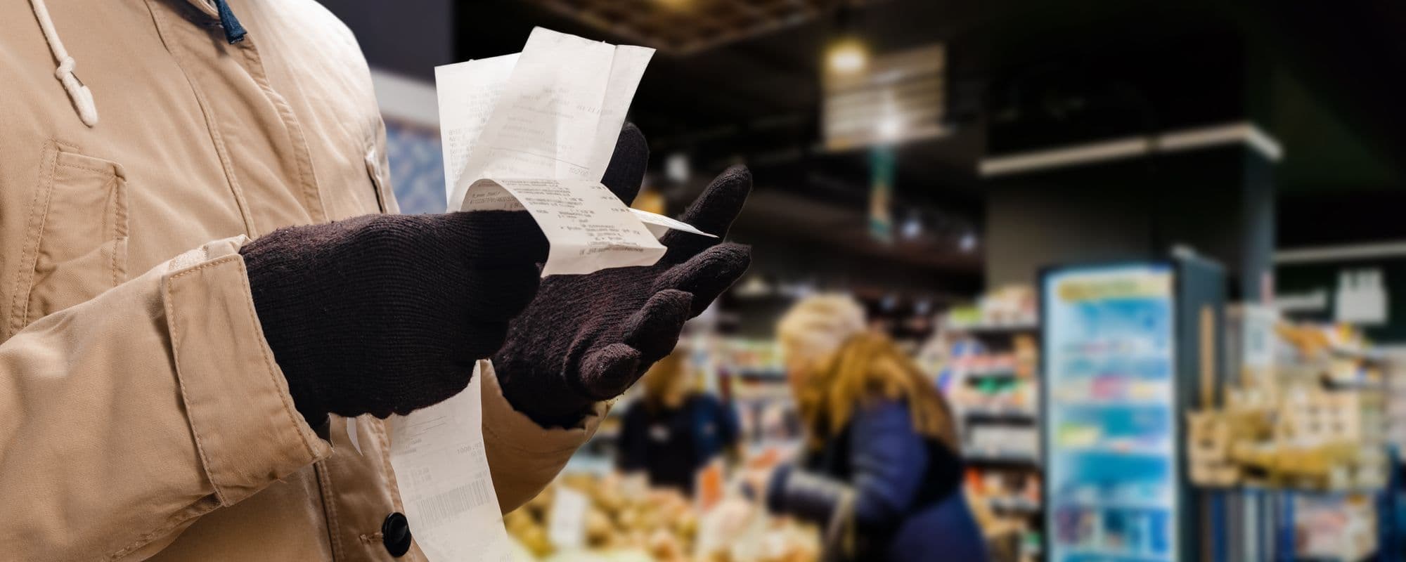Man in a jacket and gloves looking at receipts in a supermarket, with shelves and shoppers in the background.